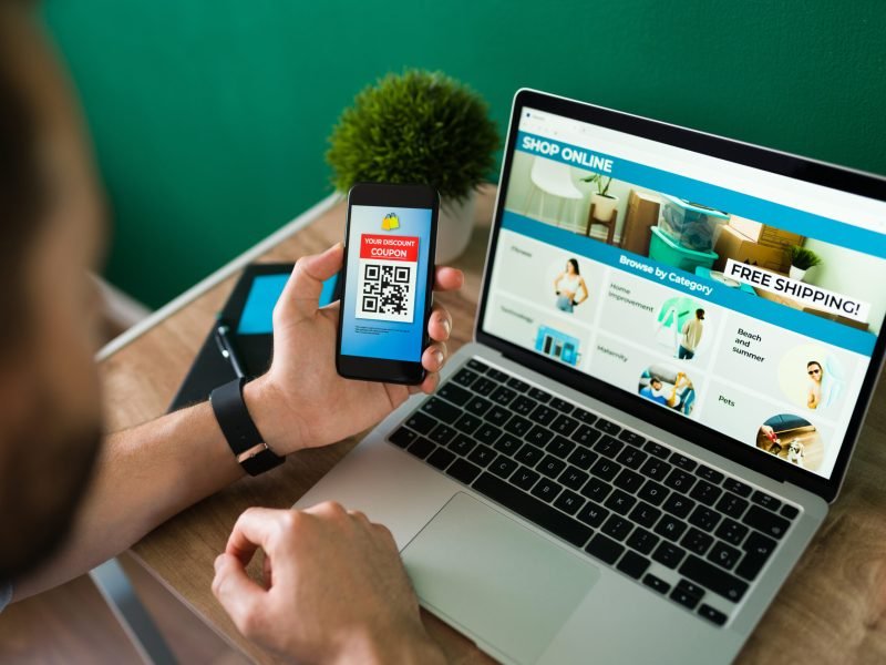 Young man using a discount coupon on his smartphone to do some online shopping on the laptop